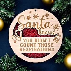 May include: A wooden Christmas ornament with the words "Santa knows... You didn't count those respirations." The ornament has a red glitter center and snowflake and medical symbol designs. Gold ornaments and green pine branches are in the background.