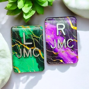 May include: Two rectangular resin tiles with a marbled design. The left tile is green with gold veins and the letters "L JMC". The right tile is purple with gold veins and the letters "R JMC".