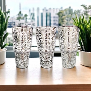 May include: Three silver tumblers with a white and silver patterned design. The tumblers are on a wooden surface.