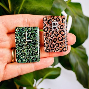 May include: Two resin letter tiles with a leopard print design. The tile on the left is green and has the letter "L" on it. The tile on the right is pink and has the letter "R" on it.