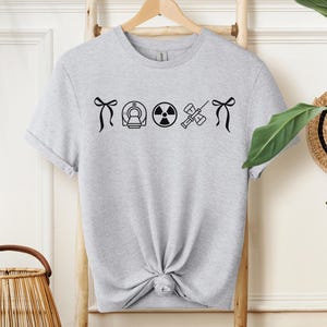 Puede incluir: Camiseta gris claro con gráficos negros de símbolos médicos, incluyendo un escáner CT, un símbolo de radiación y una jeringa. La camiseta está colgada en una percha de madera.