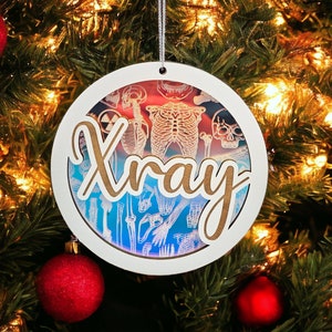 May include: A round Christmas ornament with the word "Xray" in a cursive font. The ornament features a layered design with anatomical illustrations of bones and a gradient background of red, blue, and white. The ornament hangs from a silver string.