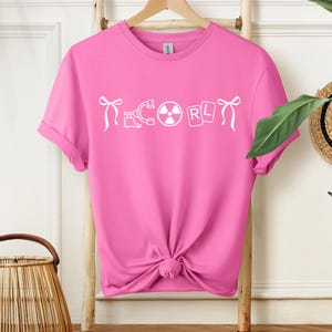 May include: A vibrant pink t-shirt featuring the word "ECORL" in white, accompanied by science-related symbols. The tee is made of a soft fabric and has a standard crew neck. The shirt is displayed on a wooden hanger.
