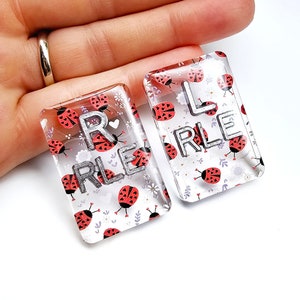 May include: Two clear rectangular resin charms with a ladybug pattern. The charms have the letters "R" and "L" with the word "RLE" written underneath each letter.