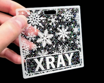 May include: White plastic name tag with a glittery snowflake design and the word "XRAY" printed in white.