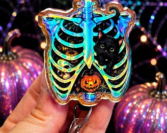 May include: A holographic badge reel featuring a human rib cage design with a black cat, a jack-o'-lantern, and spiderwebs inside. The badge reel has a clear plastic strap with a metal clip and a retractable cord mechanism for holding identification cards.