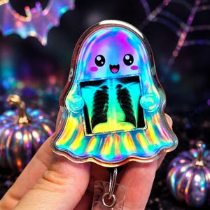 May include: A holographic ghost-shaped badge reel featuring a cute, smiling face and holding a small rectangular medical X-ray image of a human chest. The ghost has a shimmering, iridescent finish that reflects various colors of the rainbow.