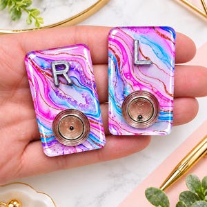 May include: Two rectangular resin pieces featuring a swirling marbled pattern in shades of pink, blue, and purple with copper glitter accents. One piece displays the silver letter R and the other the letter L, each with a circular metal component.