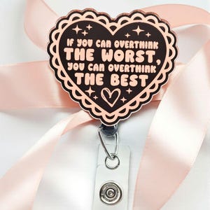May include: A rose gold heart-shaped badge reel with the quote "IF YOU CAN OVERTHINK THE WORST, YOU CAN OVERTHINK THE BEST" in white text. The badge reel has a scalloped edge and a white strap with a button. The background includes a pink ribbon.