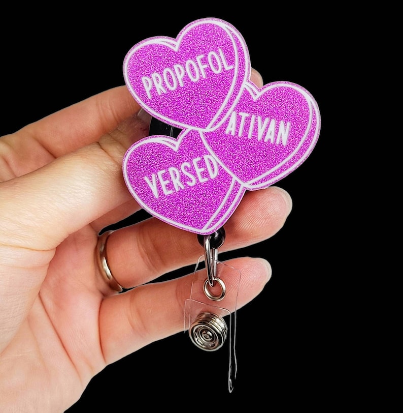 May include: A badge reel with three heart-shaped charms. Each charm is a vibrant pink with glitter and white text. The text on the charms reads "PROPOFOL", "ATIVAN", and "VERSED". The reel has a black retractable cord and a clear plastic clip.