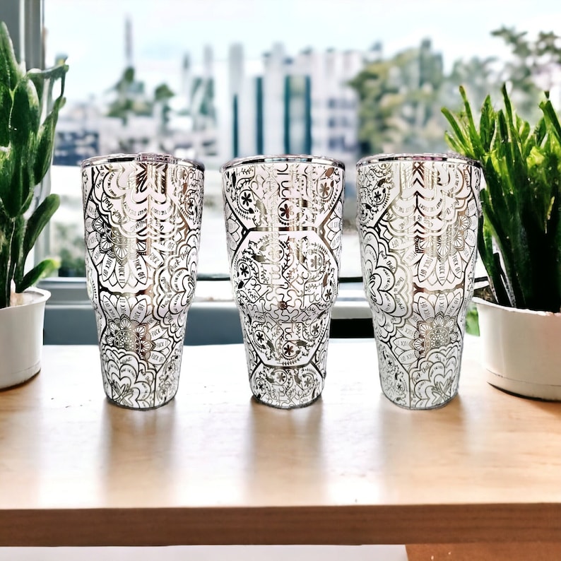 May include: Three silver tumblers with a white and silver patterned design. The tumblers are on a wooden surface.