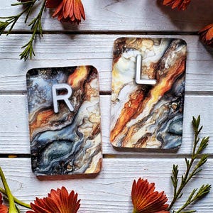 May include: Two rectangular coasters with an abstract design in shades of brown, orange, blue, and white. Each coaster has a white letter, "R" and "L", respectively. The coasters are on a white wooden surface, with orange flowers and green stems.