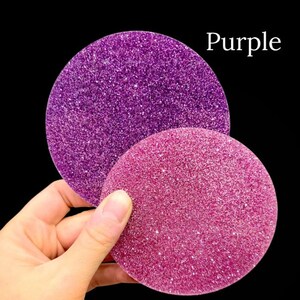 May include: The image displays two circular glitter samples against a black background. One is a vibrant purple, and the other is a soft baby pink. The text "Glitter Colors" and the color names are visible. The samples are held by a hand.