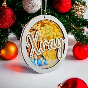 May include: A round Christmas ornament with the word "Xray" in a wooden font. The ornament has a white border and a transparent center with a skeleton design. Red and gold Christmas ornaments and green pine branches are in the background.
