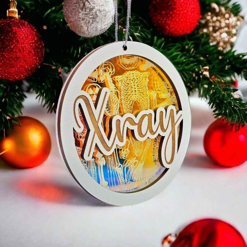 May include: A round Christmas ornament with the word "Xray" in a wooden font. The ornament has a white border and a transparent center with a skeleton design. Red and gold Christmas ornaments and green pine branches are in the background.