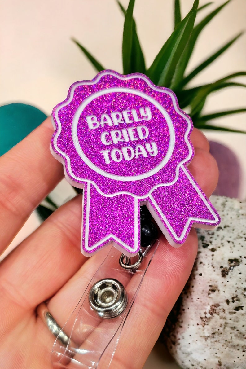 May include: A pink glitter badge holder with a white ribbon and the text "Barely Cried Today".