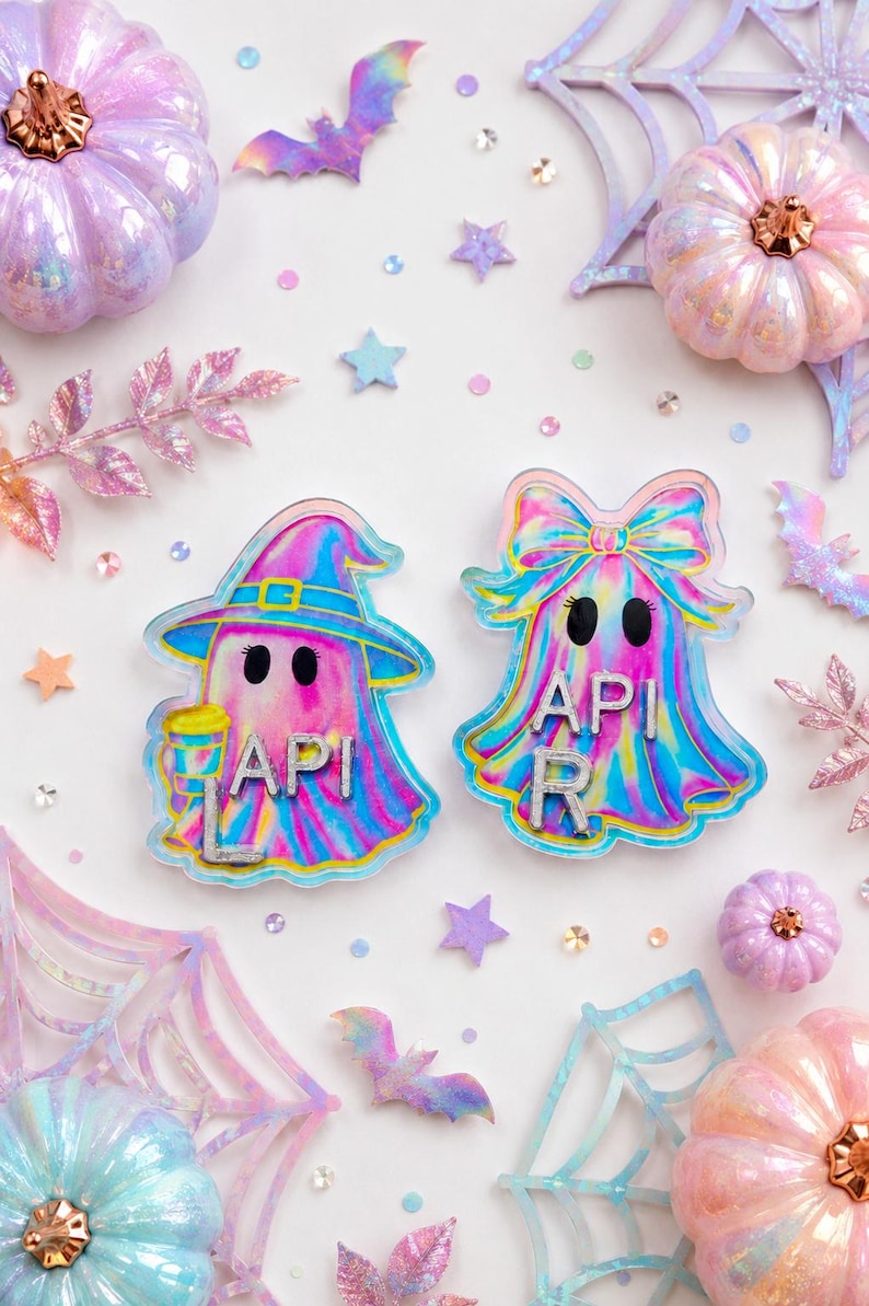 May include: Two iridescent ghost-shaped acrylic charms featuring a holographic finish. The left ghost wears a witch hat and holds a cup, with the letters A, P, I, and L. The right ghost wears a large bow, with the letters A, P, I, and R.