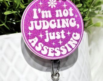 May include: A pink glitter badge holder with a white text overlay that reads "I'm not judging, just assessing".