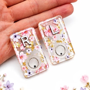 May include: Two rectangular clear resin pieces featuring embedded pink and purple floral patterns with gold leaf flakes. Each piece contains a silver letter, R or L, and a circular silver button mechanism with a small black dot in the center.