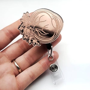 May include: Rose gold tone metal badge reel with a detailed engraved design of an eye with floral accents. The reel is attached to a clear plastic retractable badge holder.