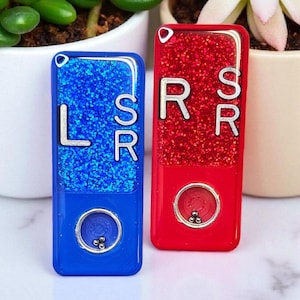 May include: Two rectangular, glitter-filled accessories, one blue and one red. The blue accessory has the letters "LSR" in silver, and the red one has "SRR". Each has a silver circle with three small silver balls.