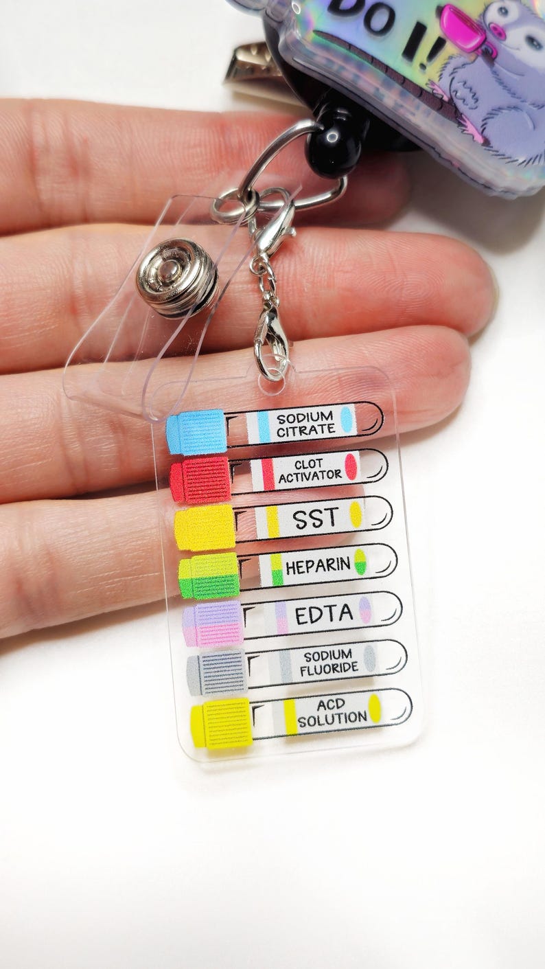 Op de afbeelding: Een doorzichtige plastic badge reel met een intrekbaar koord en een metalen clip. De badge reel heeft een ontwerp met een cartoon opossum en de woorden "DO IT!". Bevestigd is een doorzichtige plastic kaart met kleurgecodeerde bloedbuislabels.