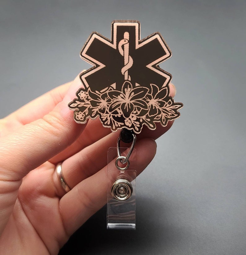 K&ouml;nnte beinhalten: Ein schwarzer und ros&eacute;goldener Lebensstern mit floralem Design, befestigt an einem einziehbaren Ausweishalter. Der Lebensstern ist ein Symbol, das von Rettungsdiensten verwendet wird.