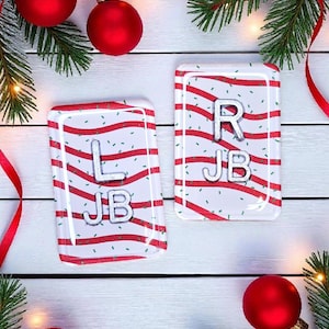 May include: Two rectangular, white and red striped Christmas ornaments with silver letters and the initials "L JB" and "R JB". The ornaments are decorated with red and green accents, and are surrounded by red ornaments, ribbons, and pine branches.