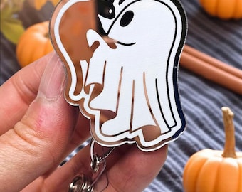 May include: A Halloween-themed badge reel featuring a white ghost and a brown cat with a black cat head. The badge reel has a silver clip and a retractable cord. The background includes small pumpkins and cinnamon sticks.