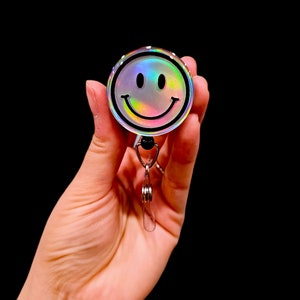 May include: A holographic iridescent silver smiley face badge reel with a black outline and a silver swivel clip.