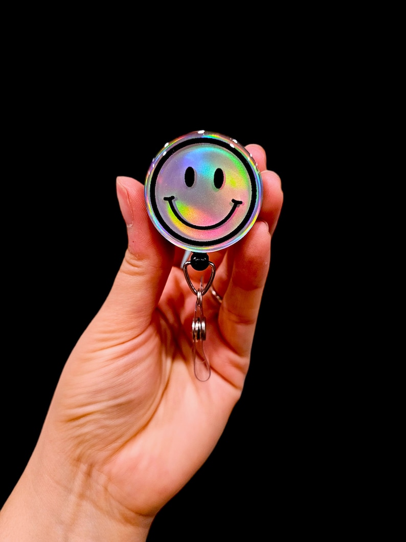 May include: A holographic iridescent silver smiley face badge reel with a black outline and a silver swivel clip.