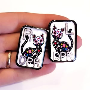 May include: Two black enamel pins with white backgrounds featuring a Day of the Dead sugar skull cat design. The left pin has the letter "R" and the text "DPR" on it. The right pin has the letter "L" and the text "DPR" on it.