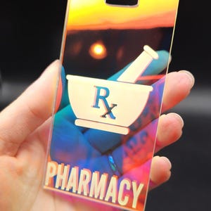 May include: A clear iridescent plastic tag with a white pharmacy symbol and the word "PHARMACY" printed in white.