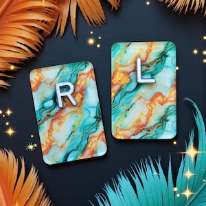 May include: Two rectangular coasters with the letters "R" and "L" in white. The coasters have a marbled design in shades of teal, orange, and gold. The background is black with orange and teal feathers and sparkling accents.