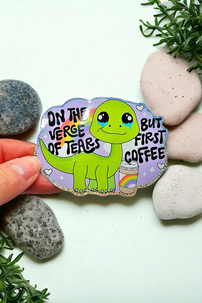 K&ouml;nnte beinhalten: Ein gr&uuml;ner Dinosaurier-Cartoon-Aufkleber mit einer Regenbogen-Kaffeetasse und dem Text "On the verge of tears but first coffee".