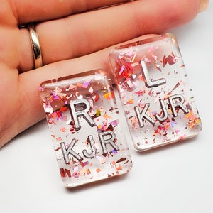 May include: Two rectangular resin pendants with silver letters spelling "R KJR" and "L KJR". The clear resin is filled with iridescent pink and red glitter. The pendants are held in a hand.