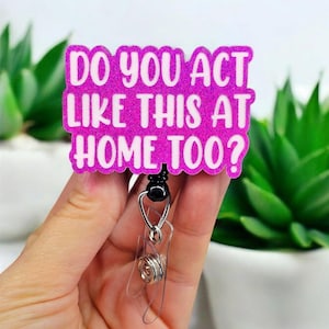 May include: A glittery pink badge reel with the text "DO YOU ACT LIKE THIS AT HOME TOO?" in white. The badge reel has a clear plastic strap and a silver metal clip. The background includes green plants in white pots.