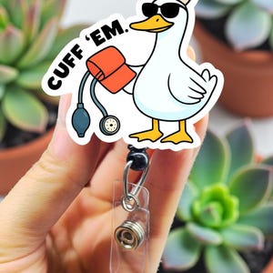May include: A white badge reel with a cartoon duck wearing sunglasses and a nurse's hat, holding a blood pressure cuff. The text "CUFF 'EM" is above the duck. The reel is held by a hand.