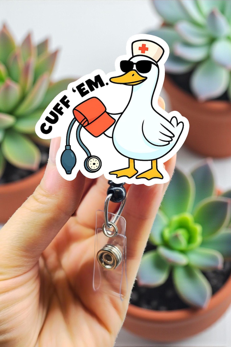 May include: A white badge reel with a cartoon duck wearing sunglasses and a nurse's hat, holding a blood pressure cuff. The text "CUFF 'EM" is above the duck. The reel is held by a hand.