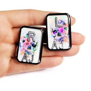 May include: Two black framed rectangular resin charms with a white background and a skull design. The skull is surrounded by colorful flowers and butterflies. The charms have the letters "RAJ" and "LAJ" printed on them.