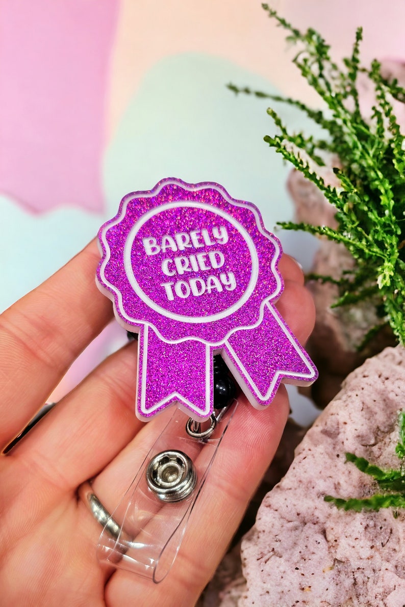May include: A pink glitter badge holder with a white ribbon and the text "Barely Cried Today".