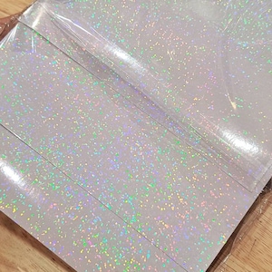 May include: A stack of iridescent white paper sheets with a holographic glitter finish. The sheets are wrapped in clear plastic.