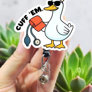 May include: A white sticker featuring a cartoon goose wearing sunglasses and a nurse's hat with a red cross. The goose holds a blood pressure cuff and the text "CUFF 'EM." is above the goose. The sticker is attached to a retractable badge reel.