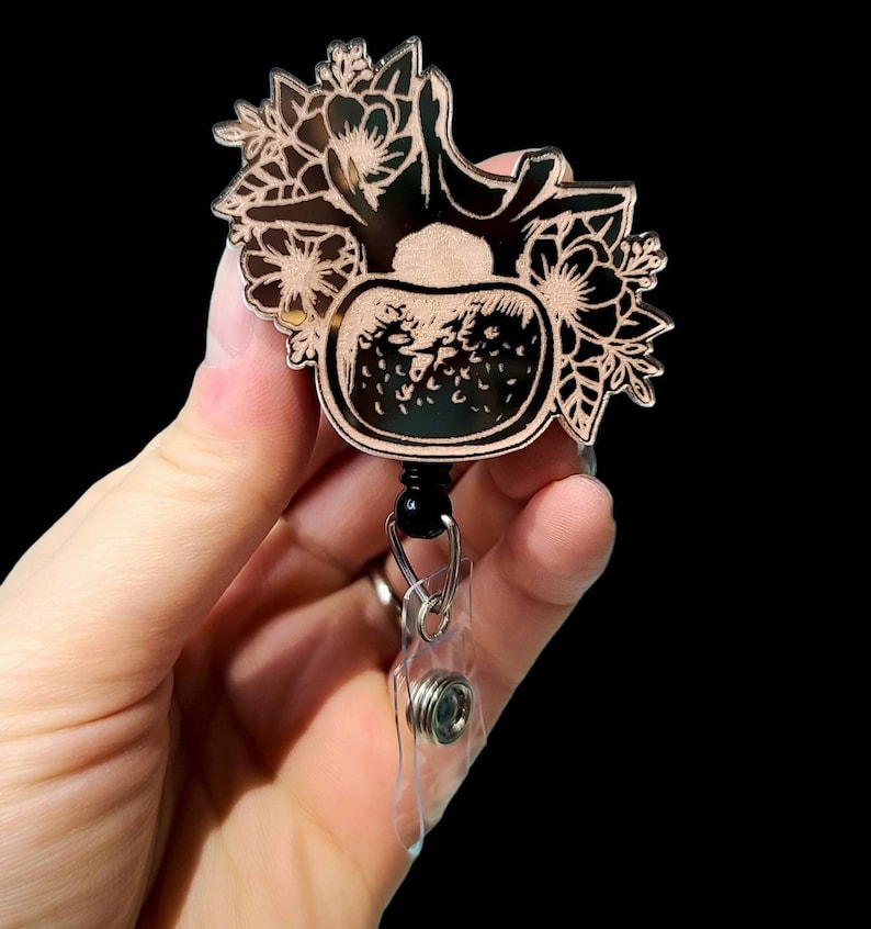 May include: A black and white floral design with a strawberry in the center, on a rose gold background. The design is attached to a clear plastic retractable badge holder.