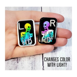 May include: Two black and white enamel pins featuring a skeleton holding a coffee cup. The pins are iridescent and change color in the light. The text "L HD" and "R HD" is on the pins.