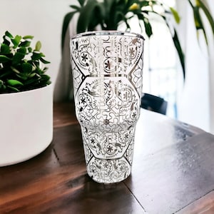 May include: A white tumbler with a silver metallic design. The design features a repeating pattern of geometric shapes and floral motifs.