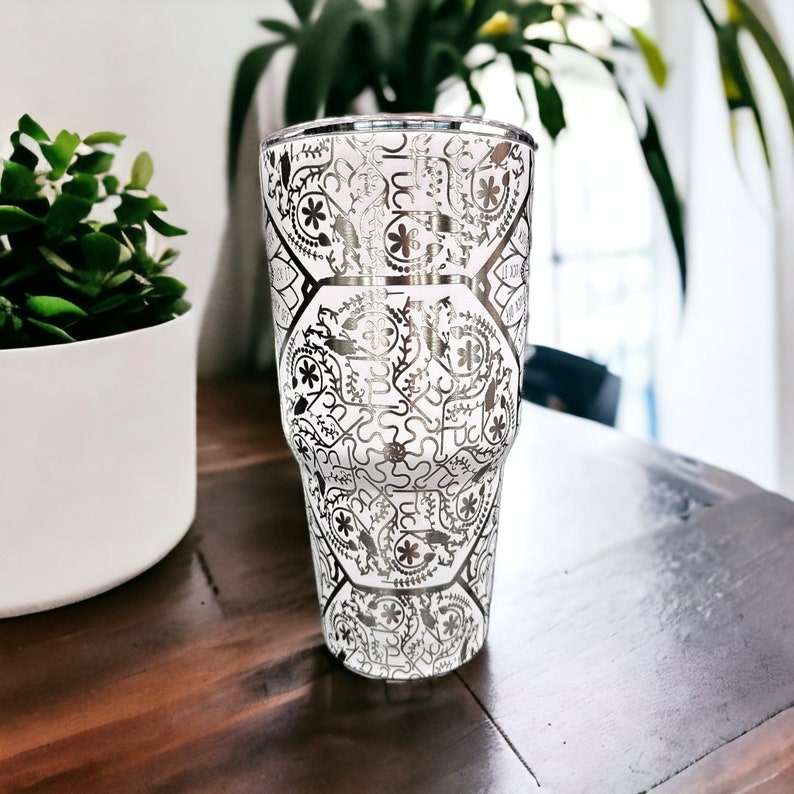 May include: A white tumbler with a silver metallic design. The design features a repeating pattern of geometric shapes and floral motifs.