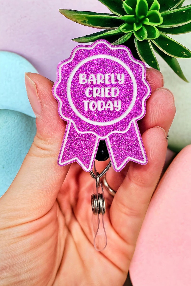May include: A pink glitter badge holder with a white ribbon and the text "Barely Cried Today".