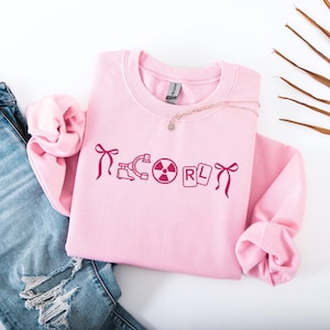 Może przedstawiać: Jasnoróżowa bluza z napisem "ICORL" w kolorze magenta, wraz z różnymi grafikami w kolorze magenta. Bluza jest zestawiona z niebieskimi dżinsami z przetarciami i delikatnym naszyjnikiem. Tło jest białe.