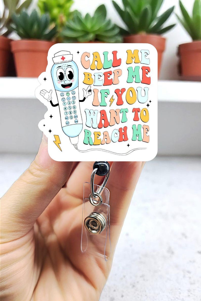 May include: A white badge reel with a cartoon telephone design. The design includes the text "CALL ME BEEP ME IF YOU WANT TO REACH ME" in colorful bubble letters. The reel has a clear plastic strap and a metal clip.
